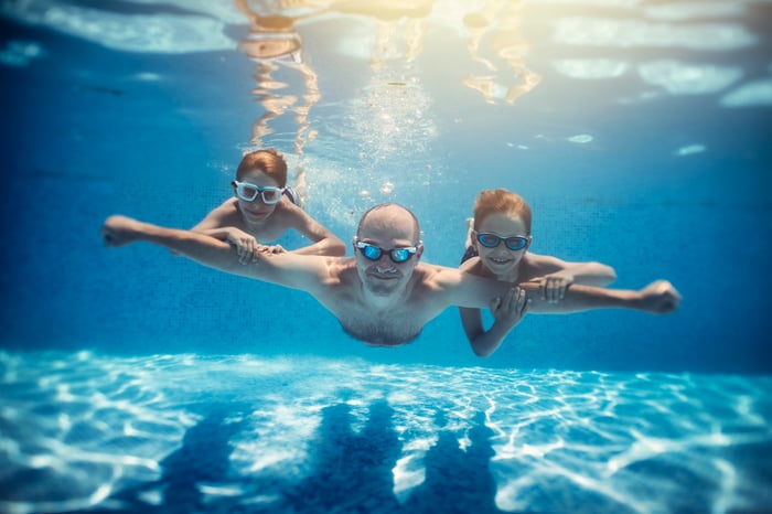Two children hold on to their parent's arms as they dive through the water in a pool.