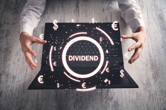 Hands near a laptop with images of currency symbols surrounding the word "Dividend" hovering over the keyboard.