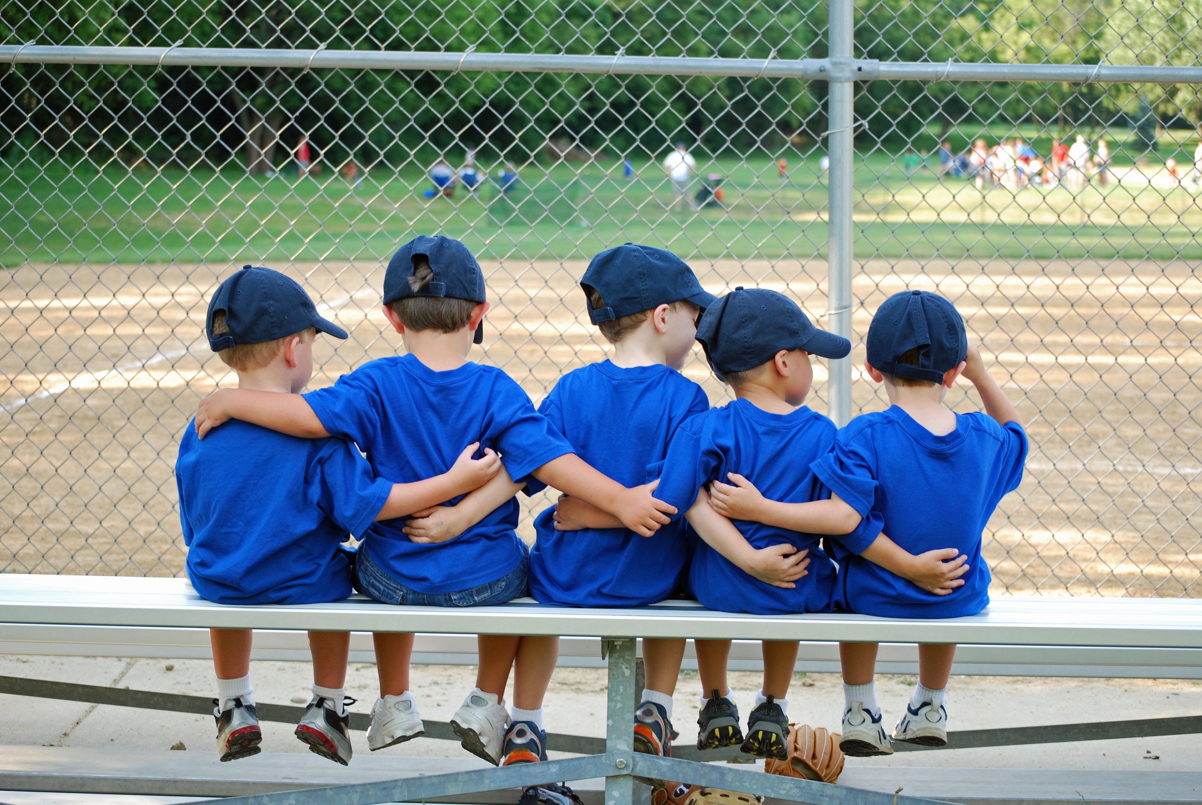 Five young baseball players wearing blue uniforms and baseball caps sit on a bench in the bleachers with their arms around each others' backs.