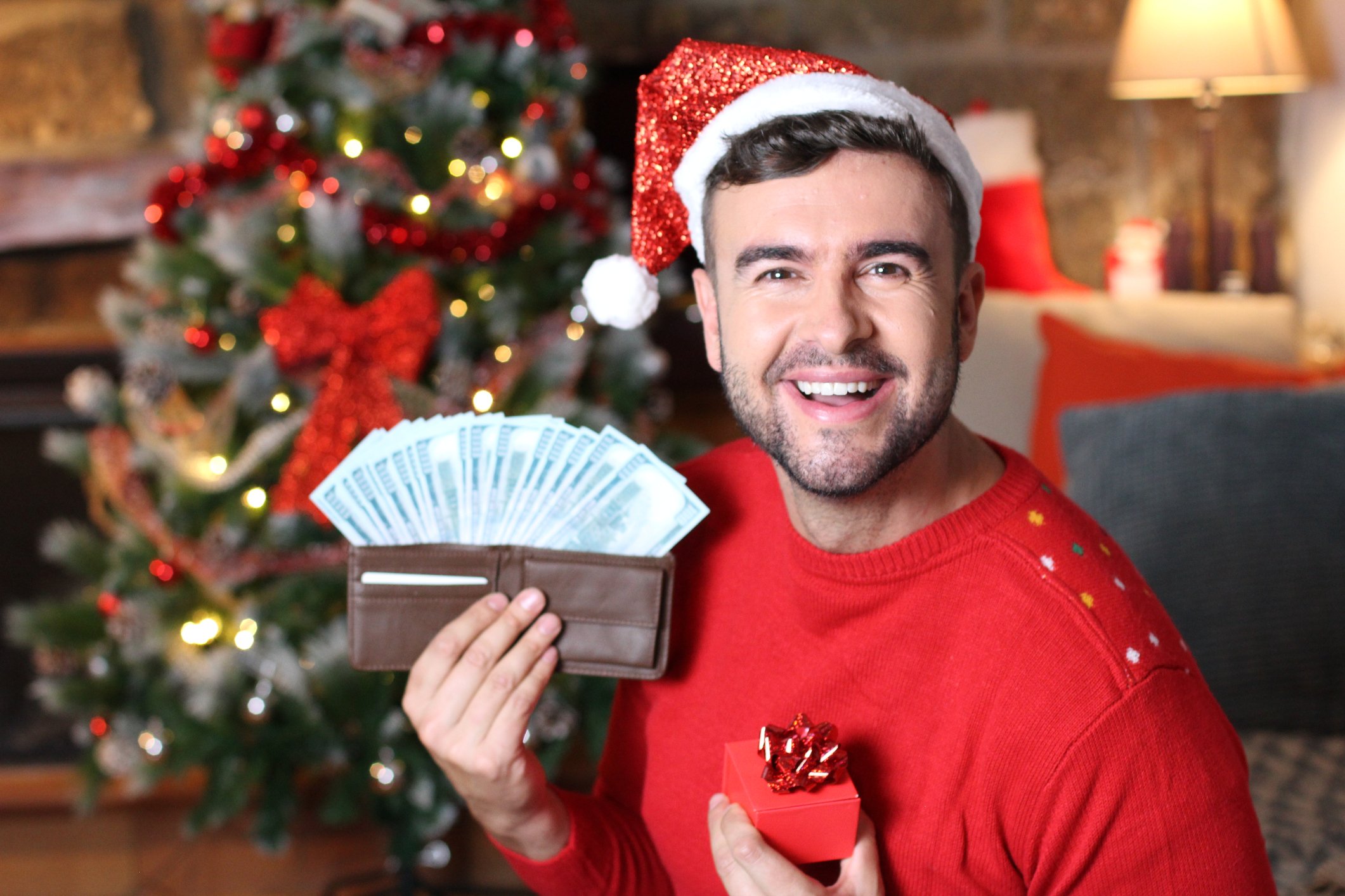 A smiling individual wearing a Christmas hat while holding a wallet filled with cash. 