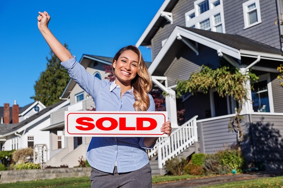 Young real estate agent with "SOLD" sign standing next to gray and white two-story home.