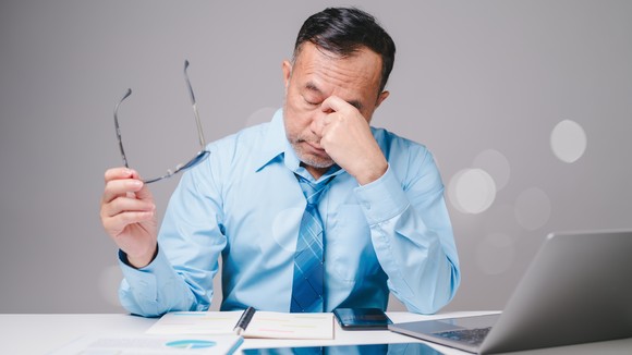 Someone seated by an open laptop has removed a pair of glasses and is frowning.