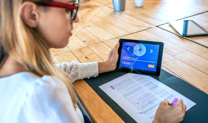 A person writing and holding a tablet displaying the logo for Ethereum.