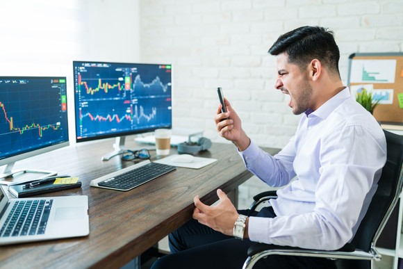 Person looking at their phone angrily while stock charts are displayed on their computer screens.