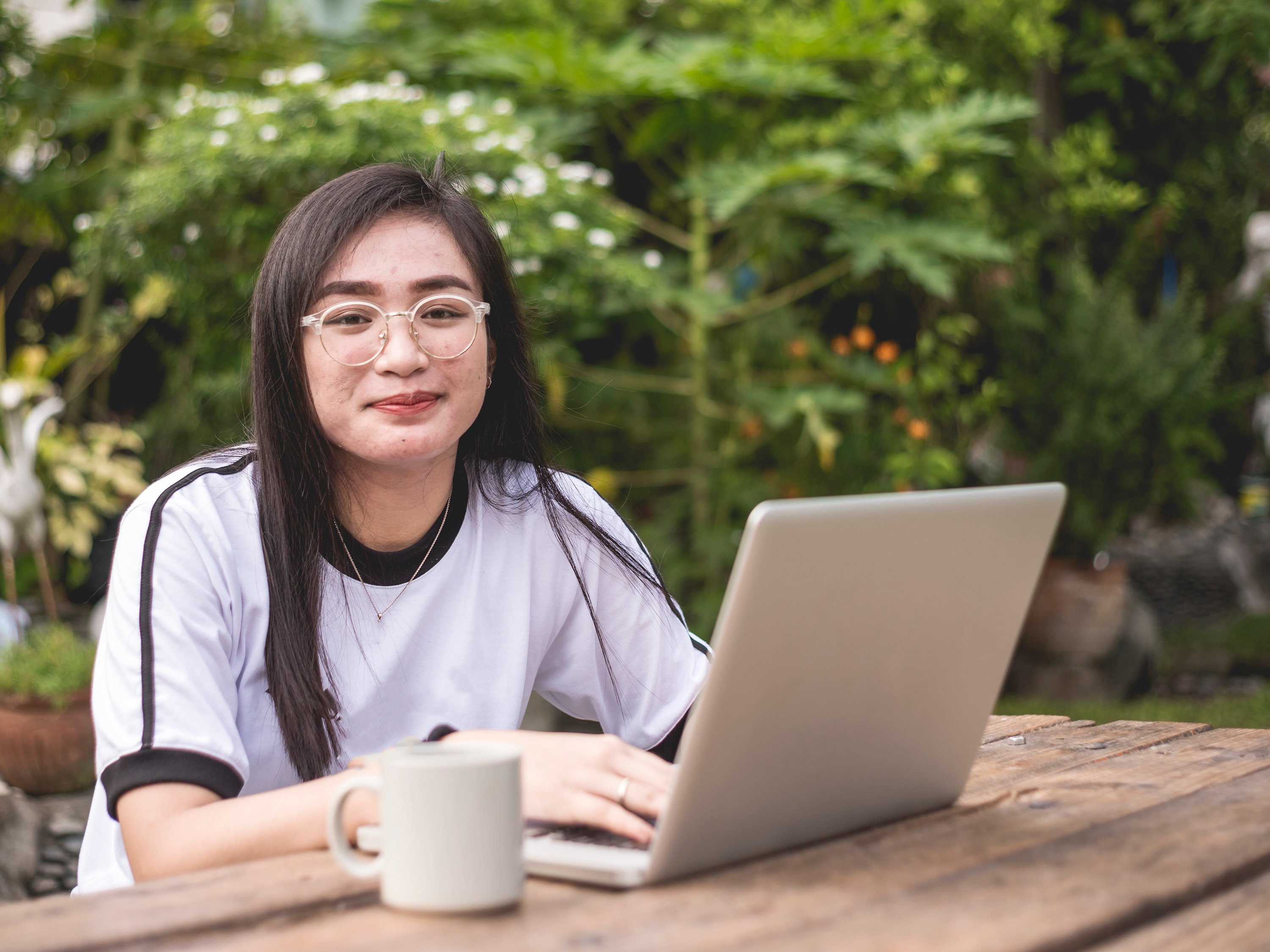 Young person sitting at a picnic table with a laptop and cup of coffee.
