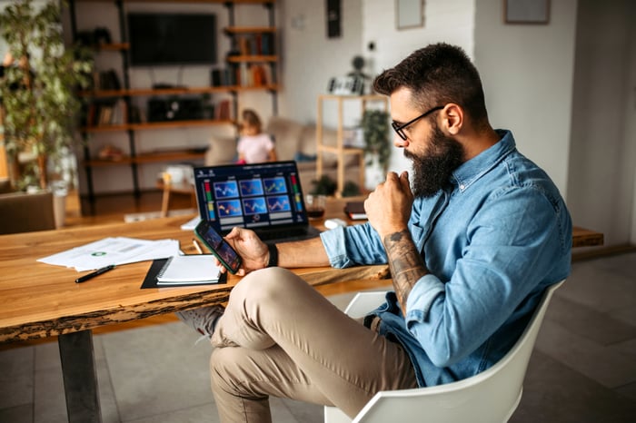 Someone looking at a smartphone while sitting at a desk in front of a laptop computer.