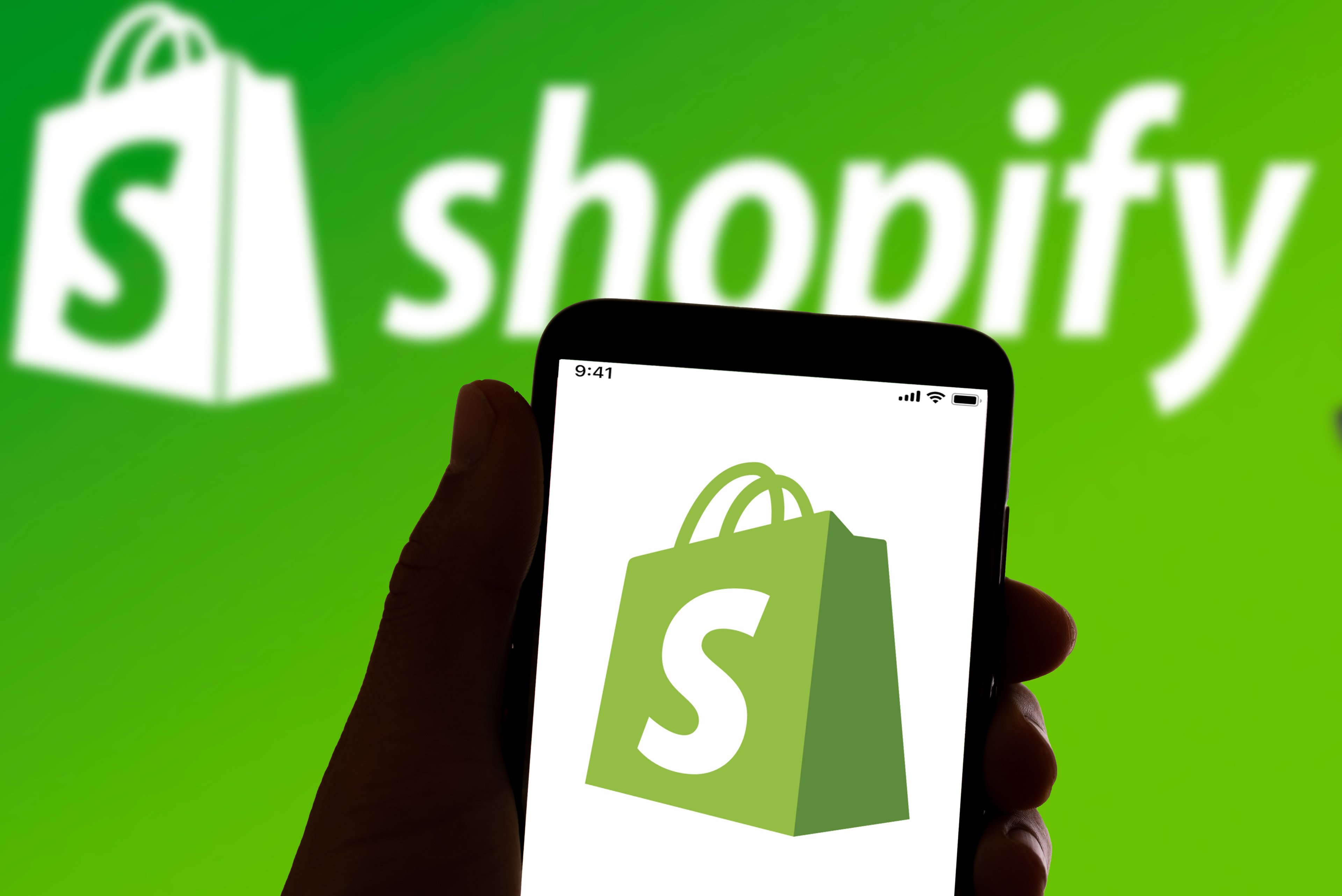 The Shopify logo of a white S on a green shopping bag shines on a smartphone that's being held up against a Shopify backdrop.