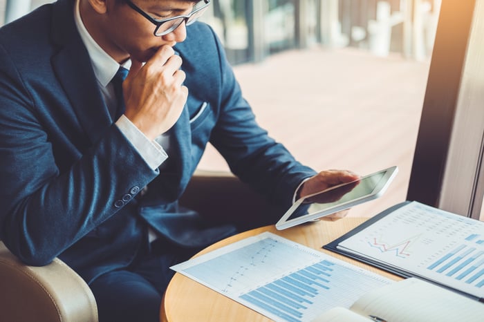A person wearing a suit looks at stock price charts on a digital tablet and paper documents.