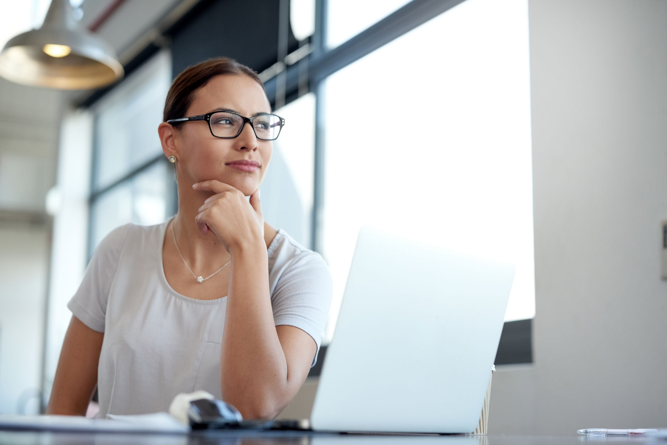 A young woman sitting in front of a laptop looking through a window into the distance. 