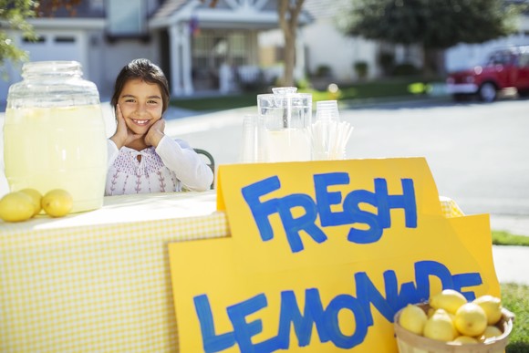 A lemonade stand on the sidewalk, with a smiling child behind the table.