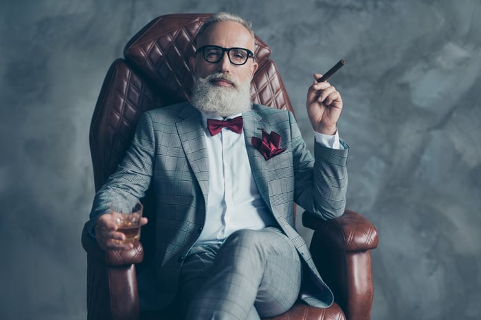 Person in a suit and bowtie smoking a cigar in a leather chair.