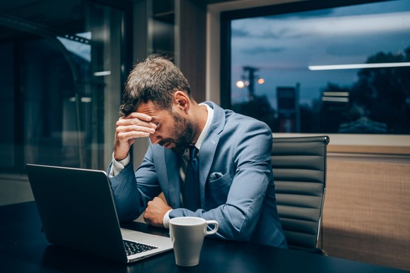Frustrated person in a business suit sitting next to a laptop, their hand to their face.