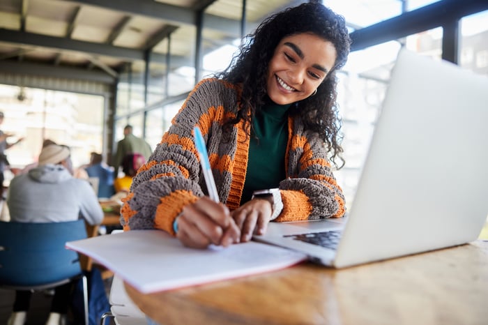 Someone writing on a paper while sitting at a desk with an open laptop.