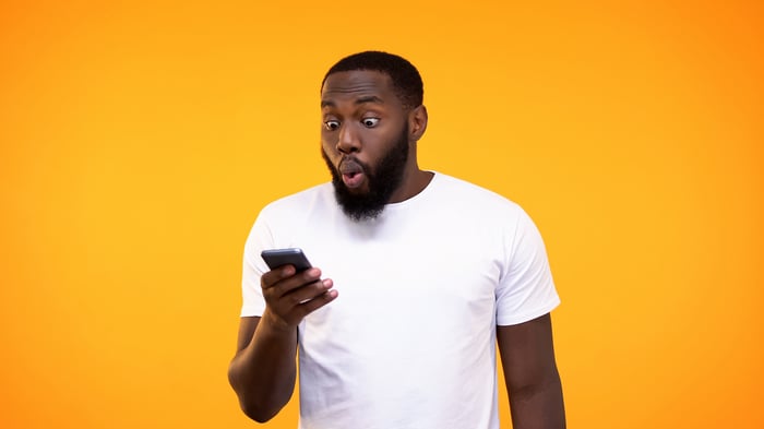 A person in a white T-shirt looking at a smartphone in shock.