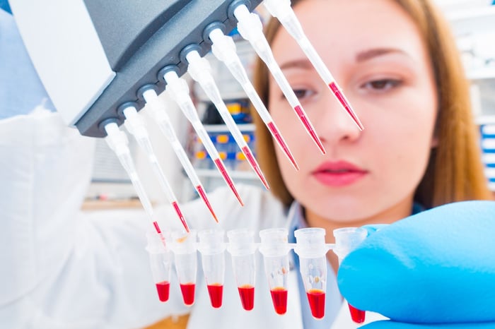 A pharmaceutical lab technician using a multi-pipette device to place red liquid into a row of test tubes. 