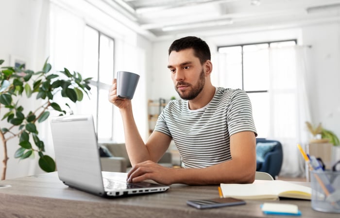 A person sitting at a desk drinking a cup of coffee while using a laptop. 