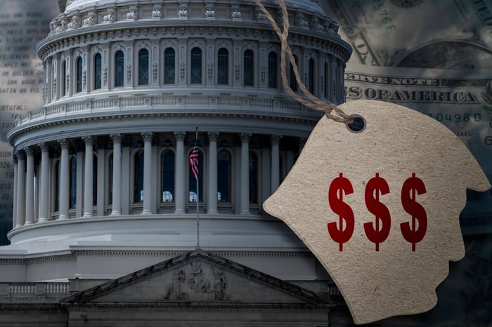 A picture of the U.S. Capitol in the background. In front  is a paper silhouette of President Trump's face with three red dollar signs printed on it. 
