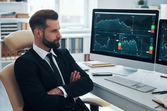 A person with arms crossed sits in front of computers showing stock prices.