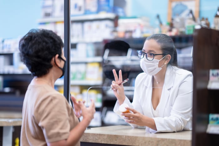 Pharmacist talking to patient through a clear divider, with both wearing masks. 