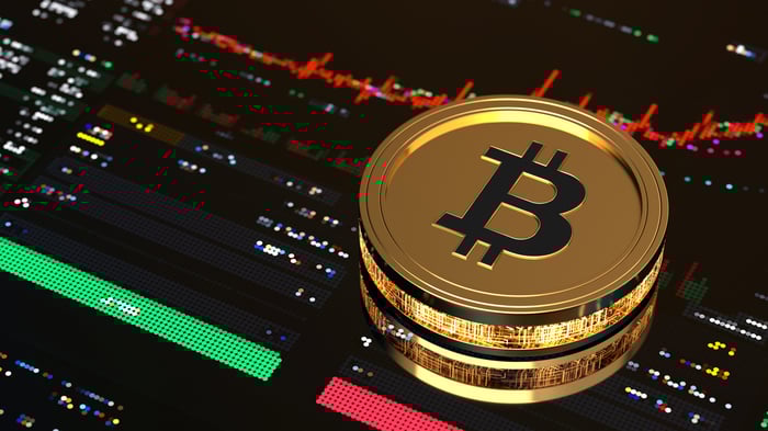 A gold coin with the Bitcoin logo sits on top of a stock chart on a tablet computer.