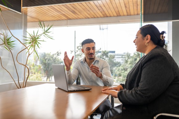 Two investors confer in a conference room as one gestures and sits in front of a laptop.
