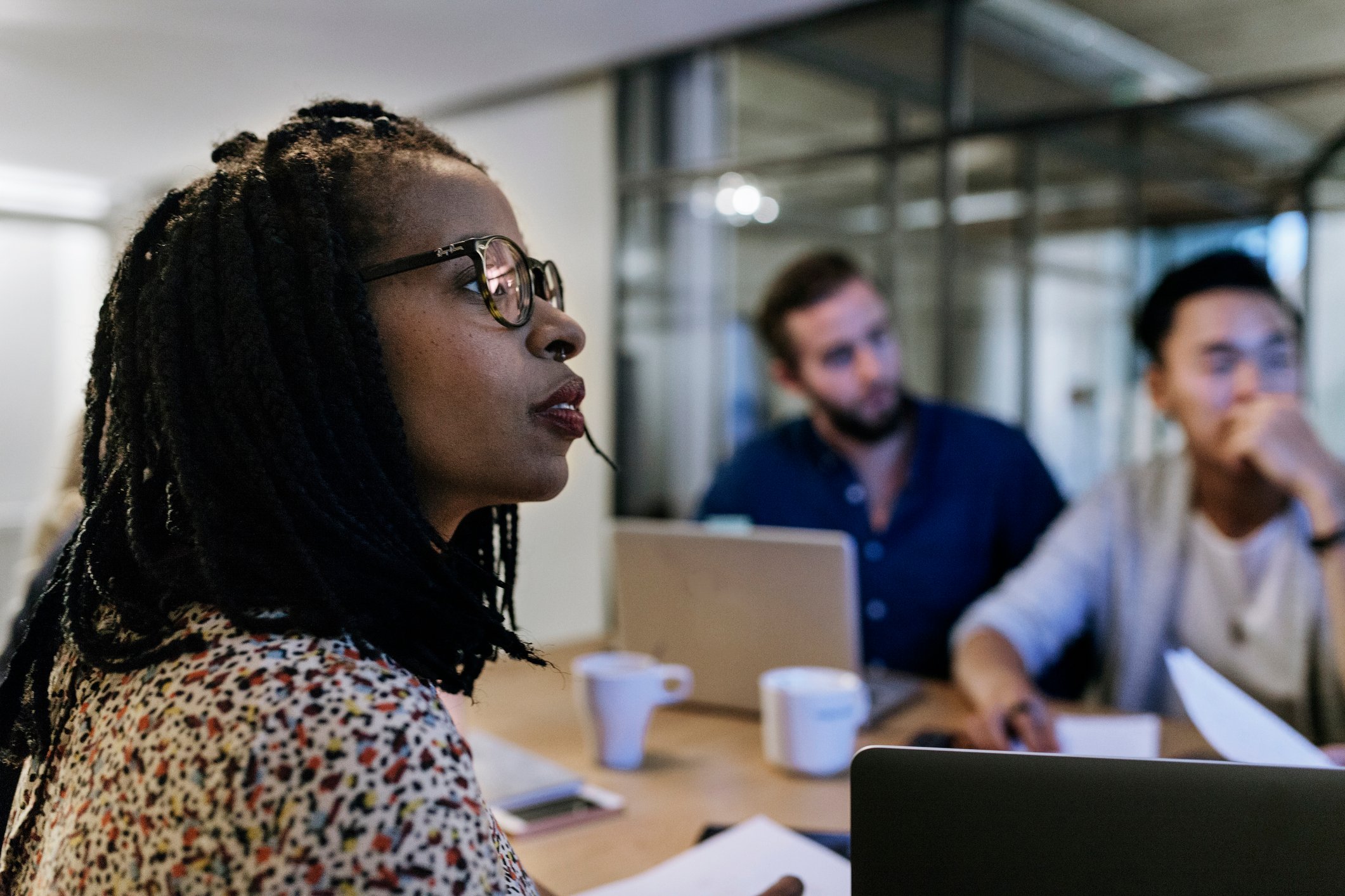 Three investors in a conference room with coffee and laptops look towards their colleagues who are making a presentation.