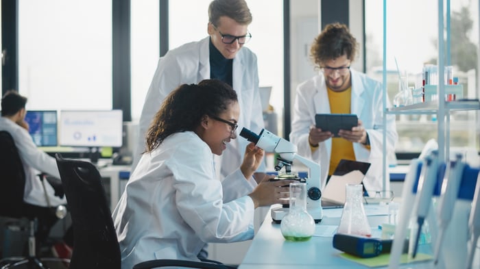 Three people wearing white coats with one looking through a microscope and another looking at a touchscreen tablet.