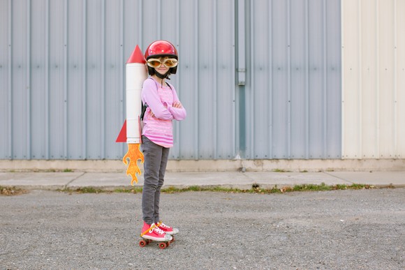 A kid wears roller skates and a toy rocket strapped to their back.