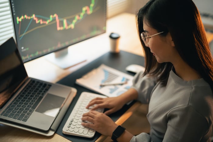 An investor types on a laptop while sitting at a desk next to a larger screen with a stock chart on it.