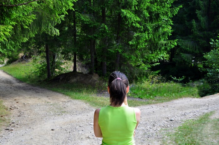 A person in a running shirt looking at a fork in an unpaved country road. 
