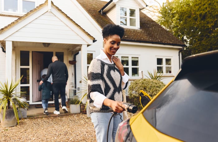 A person plugging in an electric vehicle in front of their house while other members of the family walk inside.  