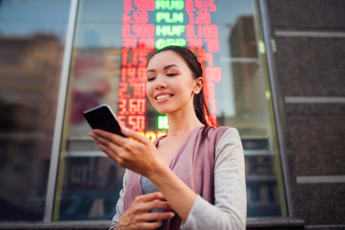 An investor stands outdoors in a city and looks at something on a phone.