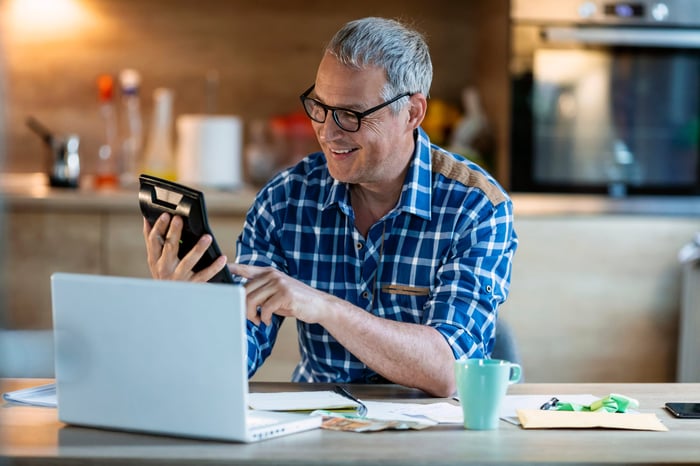 Smiling person tapping on their calculator. Their laptop is open nearby on the kitchen table.