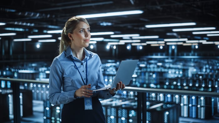 A person holding a laptop while standing in front of servers in a data center.