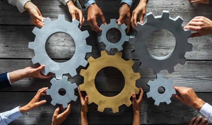 Several hands holding an assortment of gears that fit together.