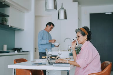 Couple in kitchen one holding mug other on laptop