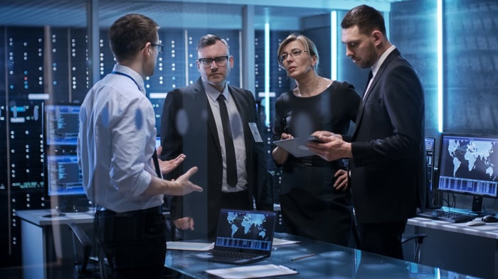A group of investors stand in a room with computers and talk while one looks at a tablet.