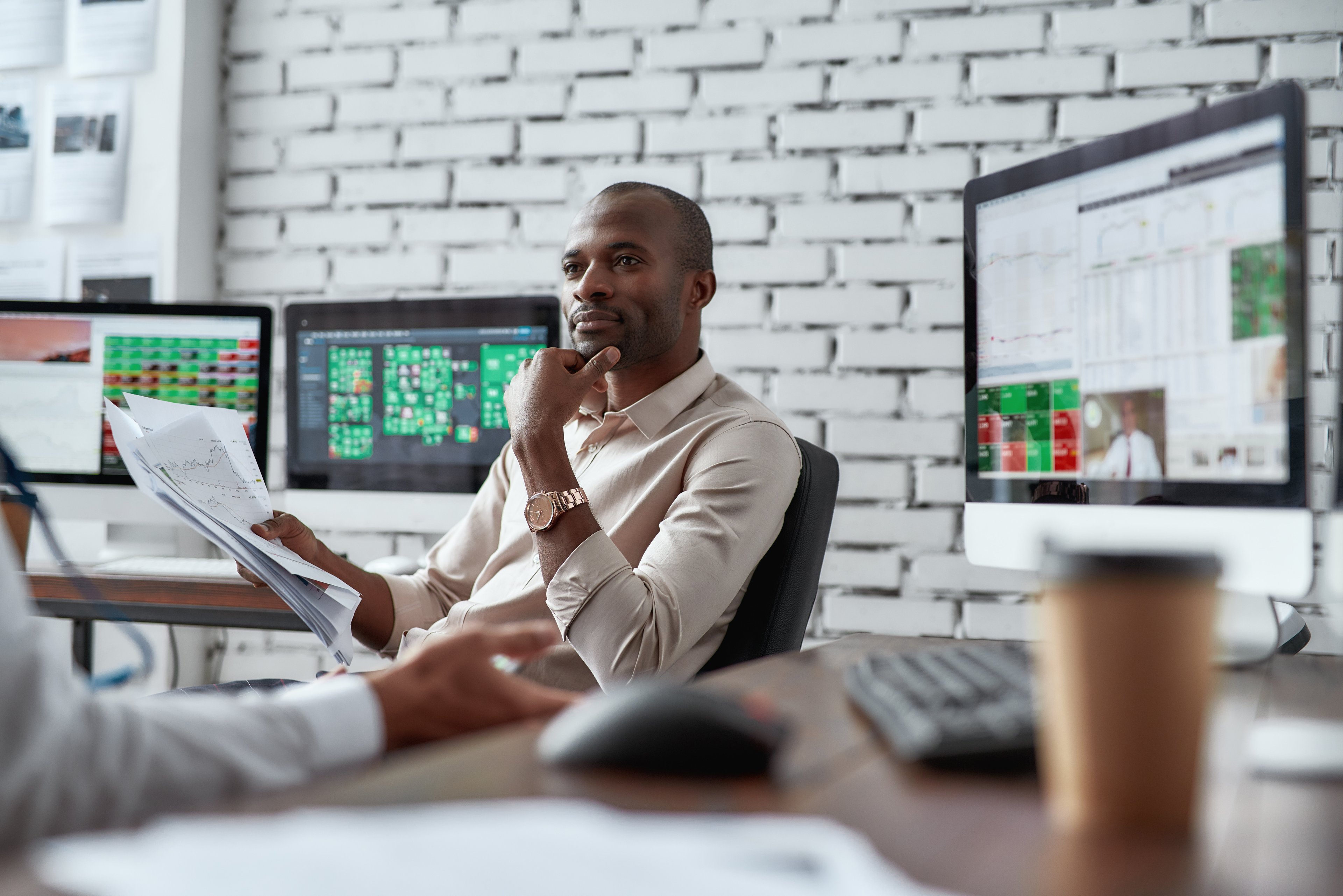 One investor talks to another at a desk in an office as screens displaying stock charts rest in the background.