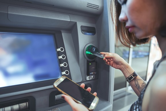 A person looking at their smartphone while inserting a card into an ATM.