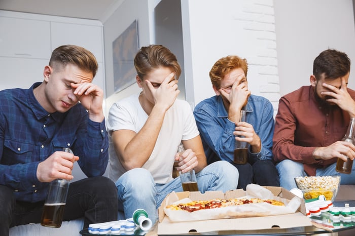 Four people sitting on a couch in front of a pizza and a bowl of popcorn touch their faces in dismay.