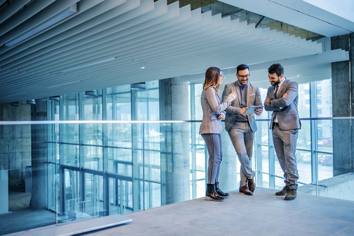 A group of three investors stand near a railing inside of an office lobby and look at a tablet computer.