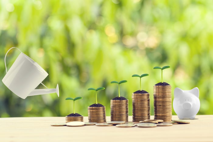 A gardner's watering can is held over ascending stacks of coins that have a sprout at the top of each of them.