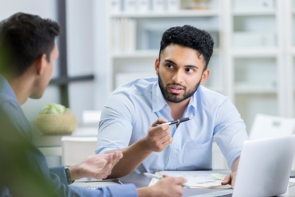 Two investors discuss figures while sitting in an office in front of a laptop and several papers.