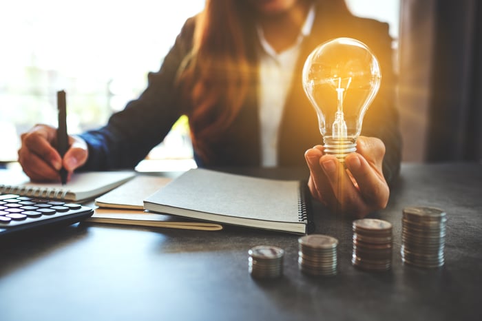 A person holding a glowing lightbulb while sitting at a table on top of which are a calculator, notebooks, and stacks of coins. 