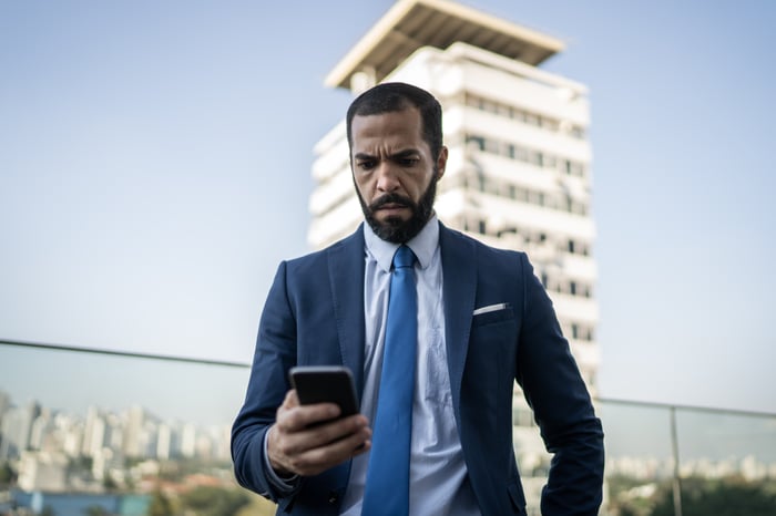 Investor in suit and tie checking smartphone amid backdrop of office tower.