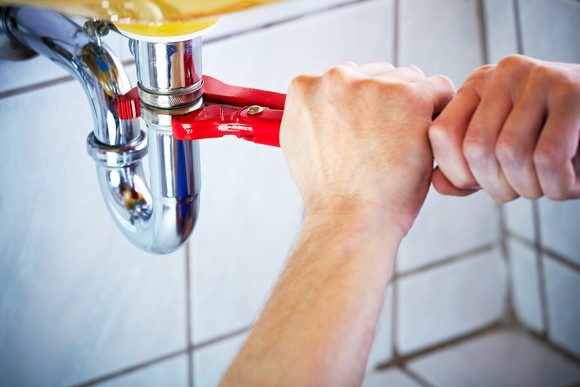 A plumber wrenches on a sink pipe, with white tiles in the background.