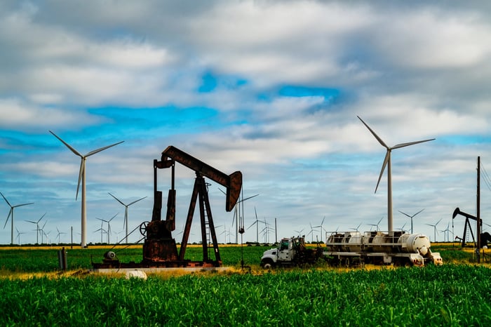 A rusty pumpjack draws up oil, while wind turbines surround it. 