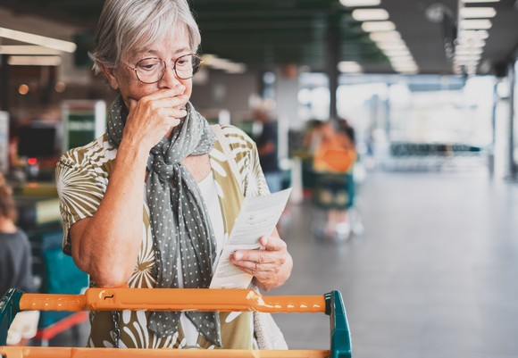 Older person stands with a shopping cart, scanning a store receipt with a worried expression on their face. 