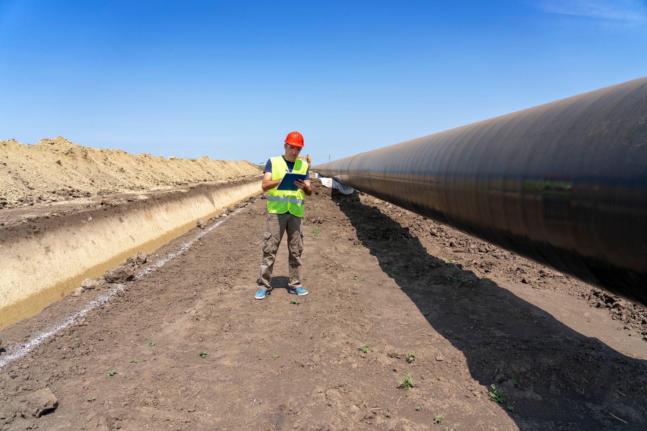 A worker wearing a hardhat looking at a touchscreen tablet while standing next to a pipeline.