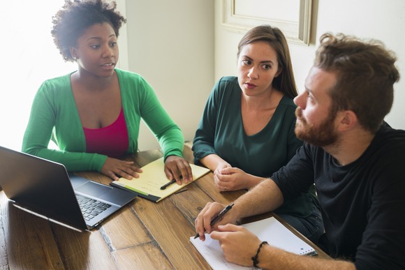 Three worried people discuss an issue while sitting at a table in front of a laptop and a couple of notepads.